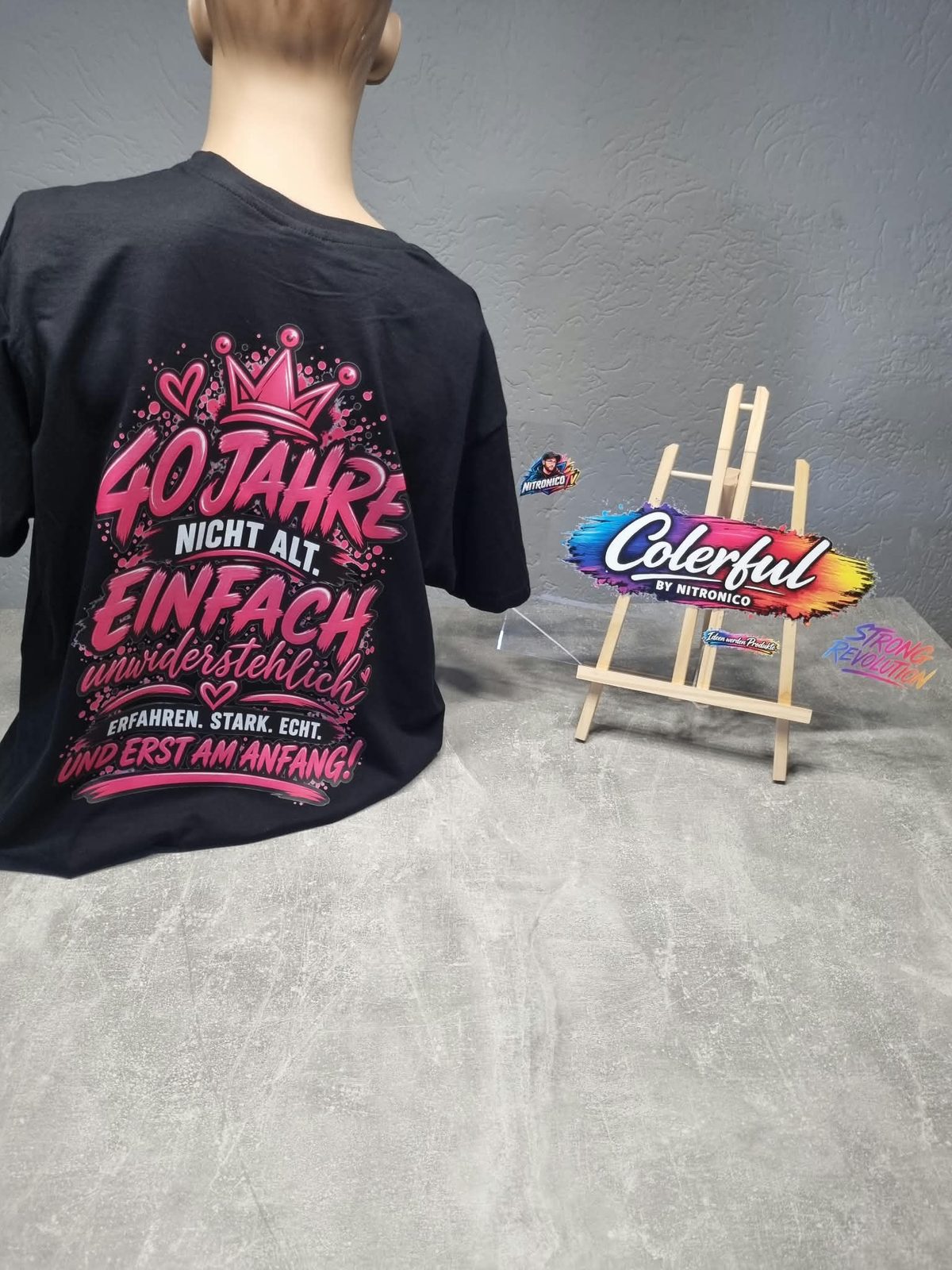 Schwarzes Shirt mit pinkem Rückenmotiv „40 Jahre nicht alt. Einfach unwiderstehlich“ neben der Colerful-Staffelei